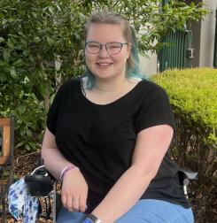 Jessi, a young woman who is a wheelchair user, wearing glasses and a black t-shirt.